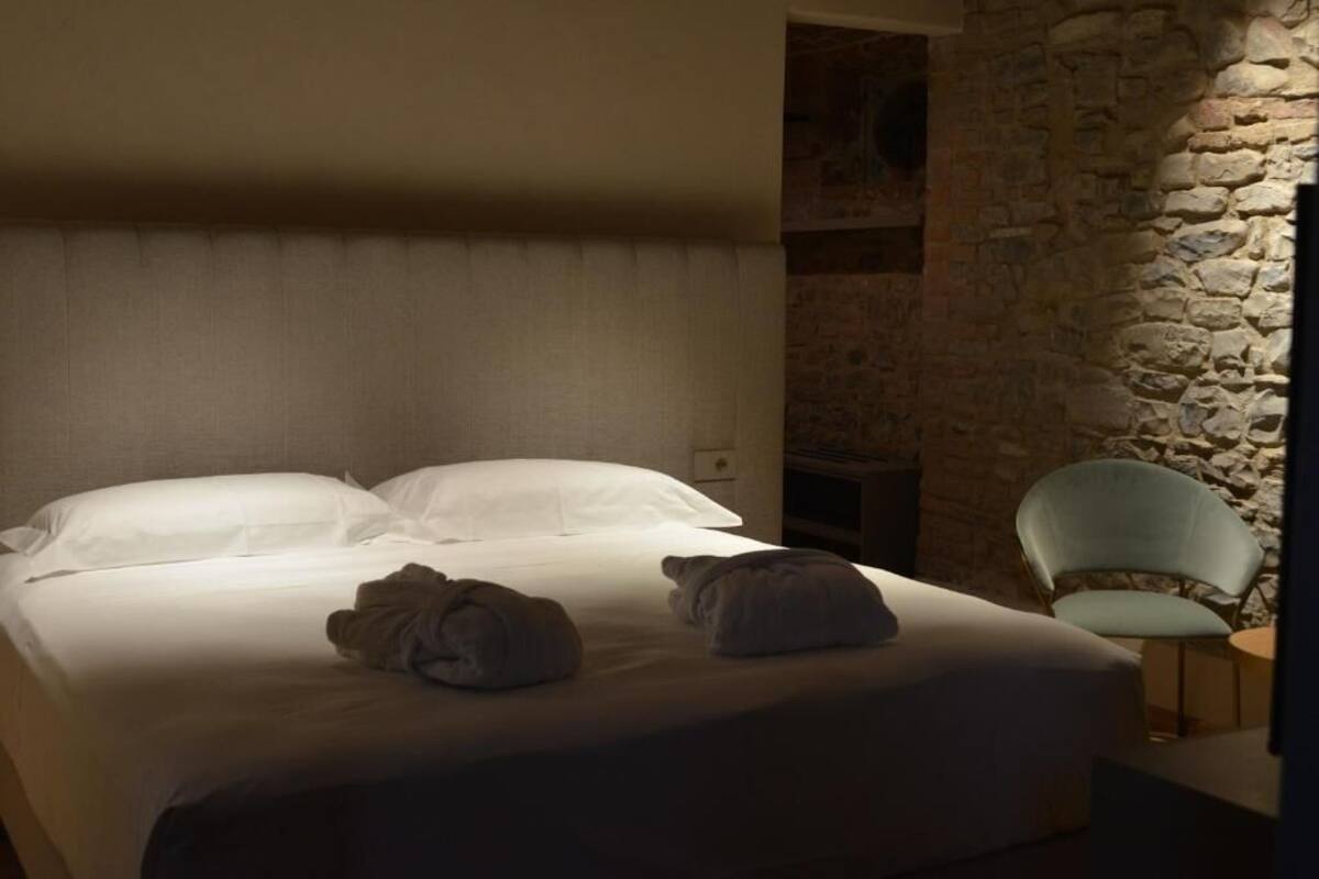 Una cama con sábanas blancas y dos toallas enrolladas, una pared de piedra y una silla verde claro.