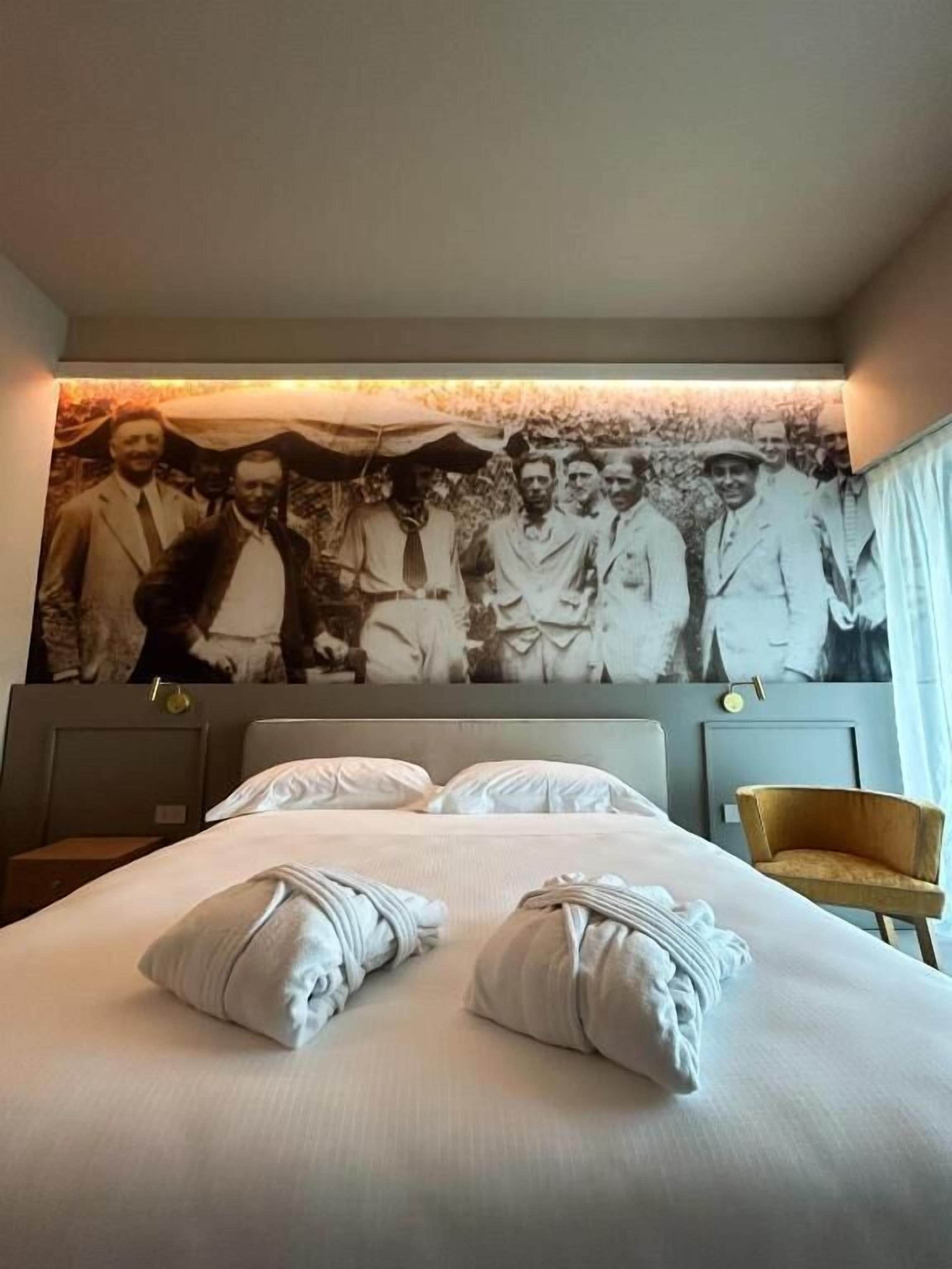 Una habitación de hotel con una cama grande, dos almohadas, una mesita de noche, una silla y una pared con una fotografía blanco y negro.