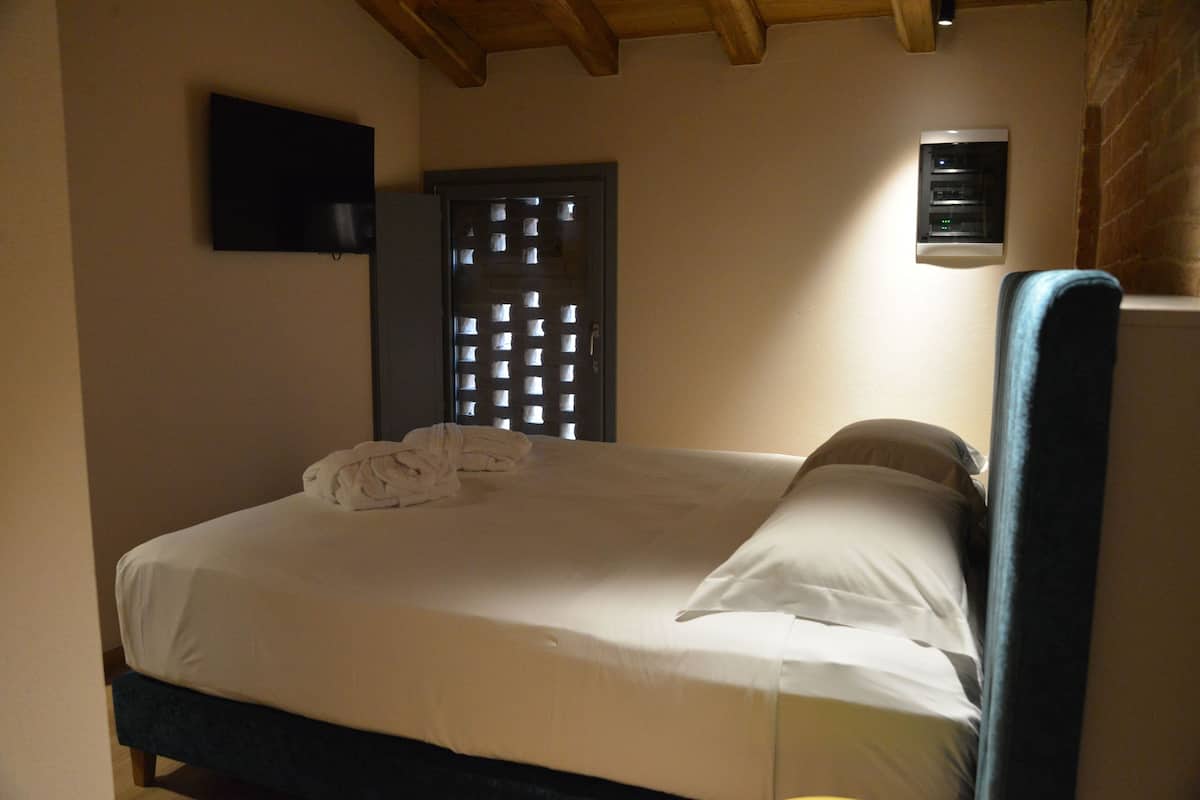 Una cama individual con colchón y almohadas blancos, cabecera oscura, televisor montado en la pared y unidad de aire acondicionado pequeña.