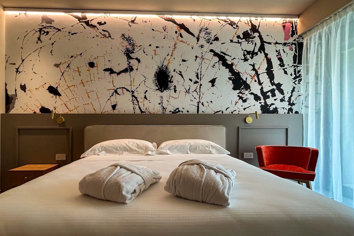 A hotel room with a large bed, two pillows, a red chair, and a wall with a unique black and white abstract design.