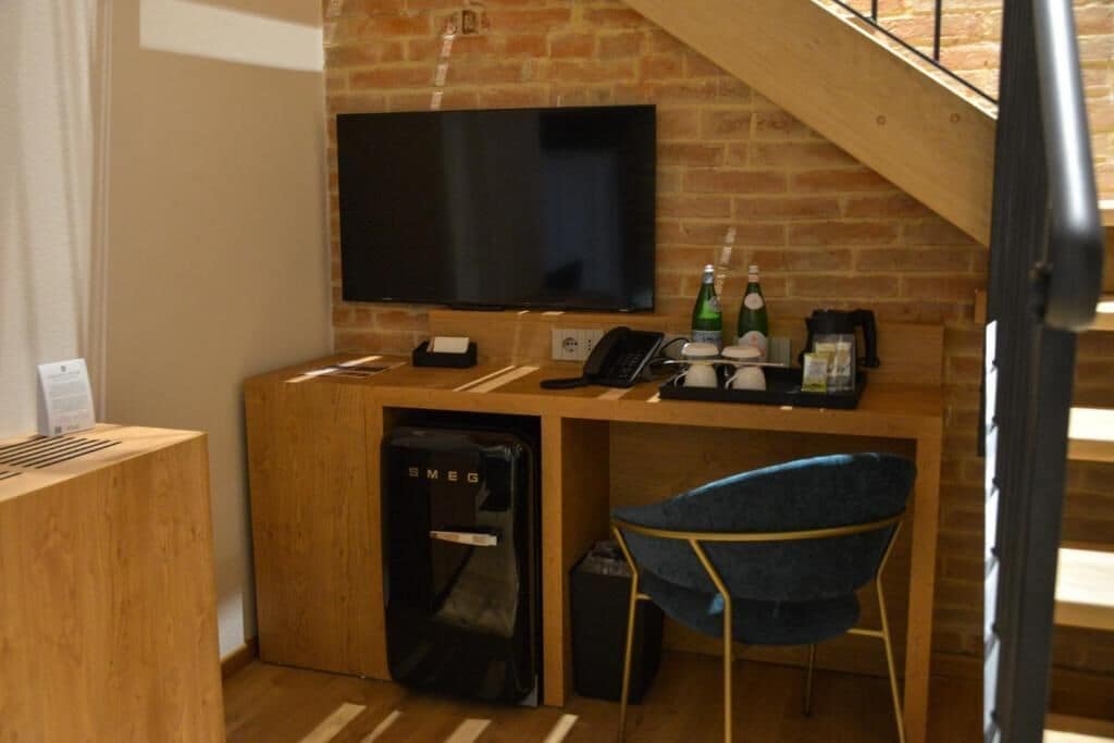 Una habitación con pared de ladrillo, un televisor de pantalla plana sobre un escritorio de madera, una silla azul y un refrigerador pequeño.