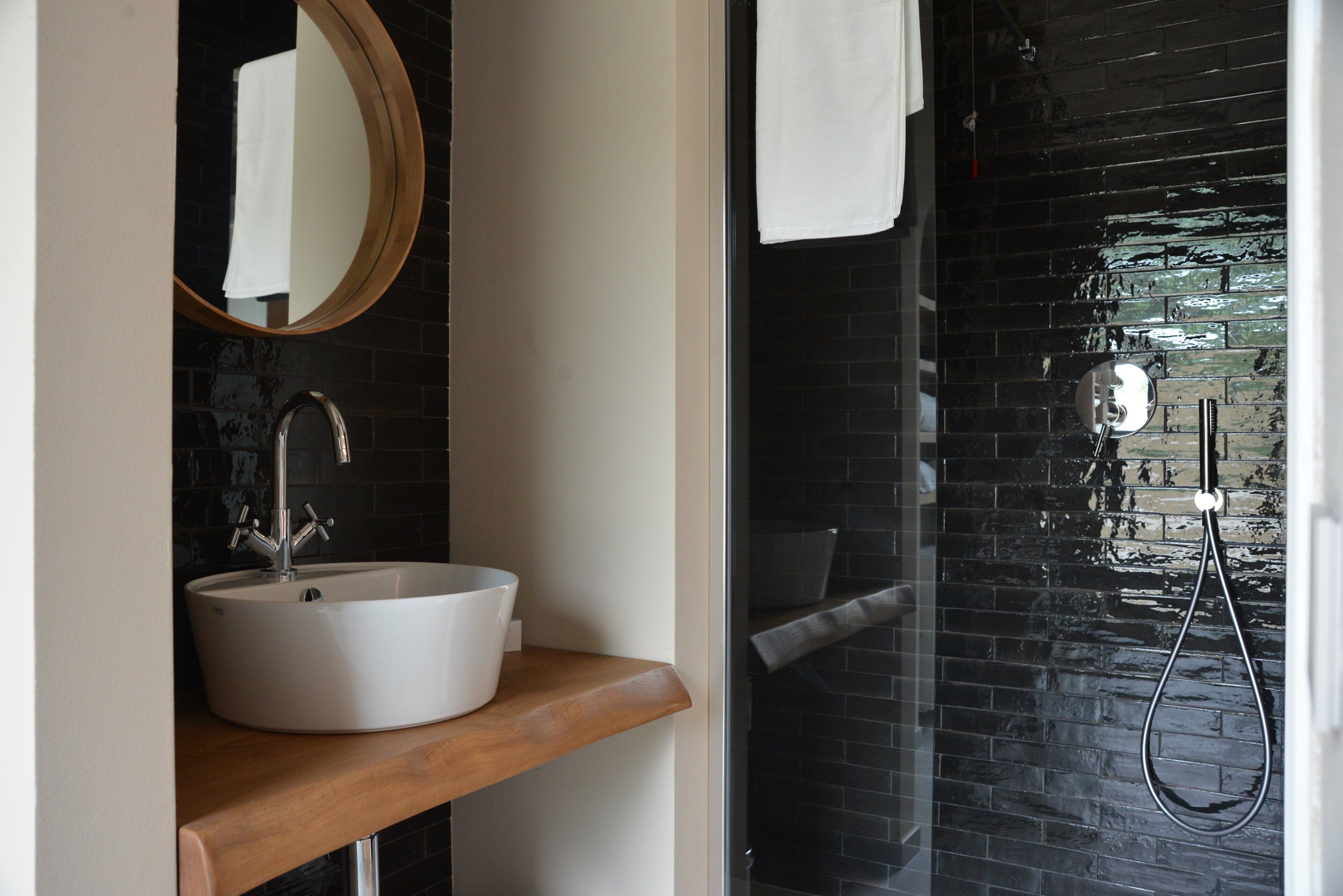 Un baño con un lavamanos blanco, una encimera de madera, un espejo redondo y una zona de ducha con azulejos negros.