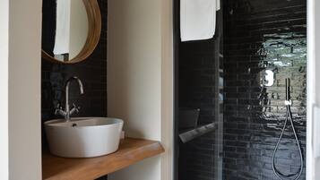 Un baño con un lavamanos blanco, una encimera de madera, un espejo redondo y una zona de ducha con azulejos negros.