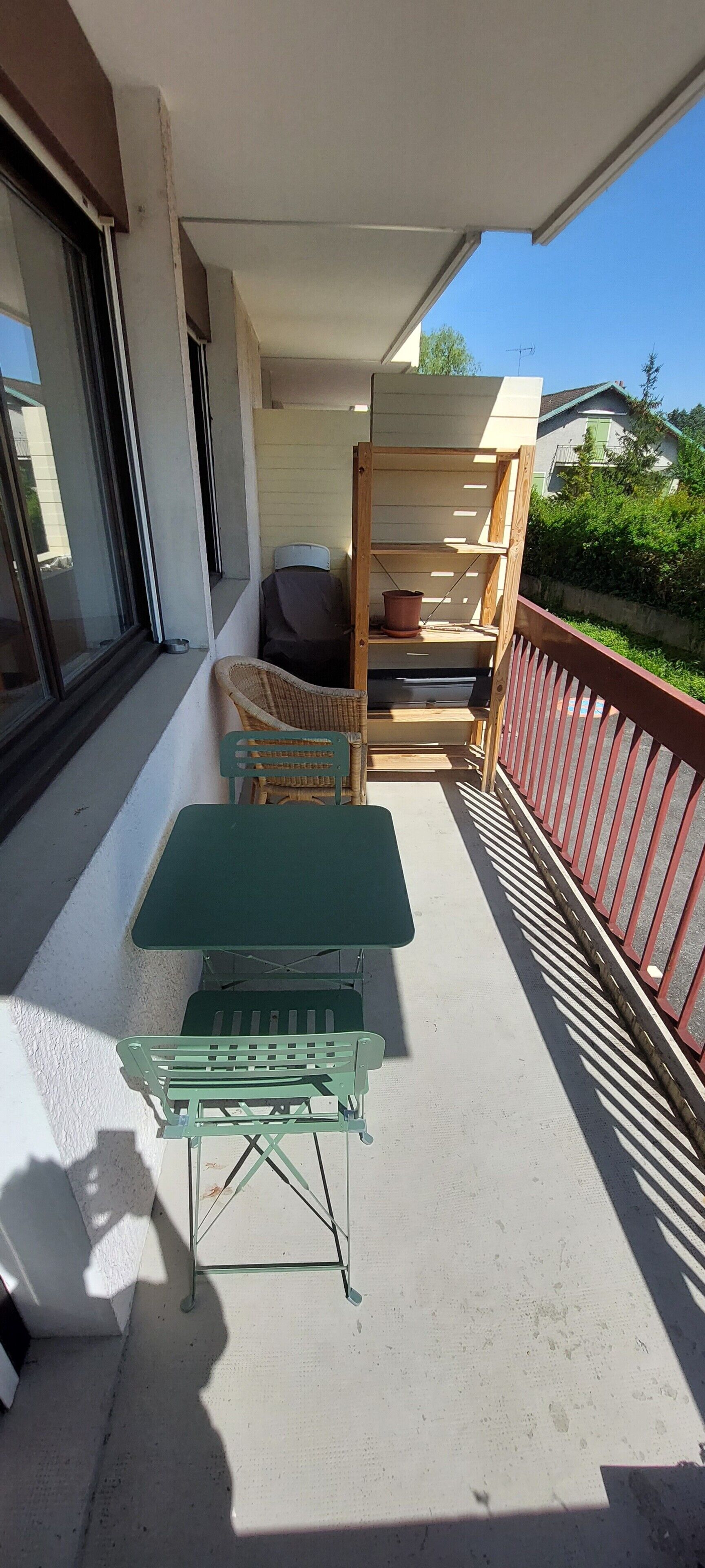 A balcony with a green table and chairs, a wooden shelf, and a red railing.