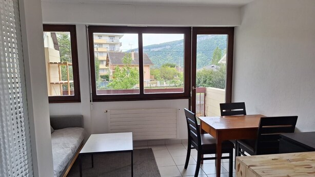 A small room with a bed, a table, and chairs, large windows with a view of buildings and trees, and a radiator. - Studio "Les Cascades" with Mountain View, Balcony, and Wi-Fi (Gaillard)
