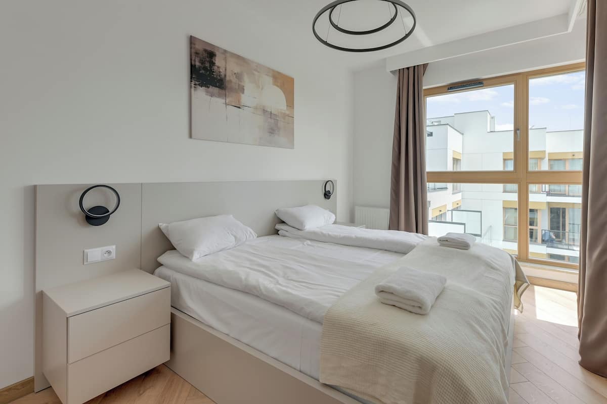 Una habitación de hotel moderna con una cama grande, una mesita de noche, una lámpara de pared y un ventanal con vistas a edificios.