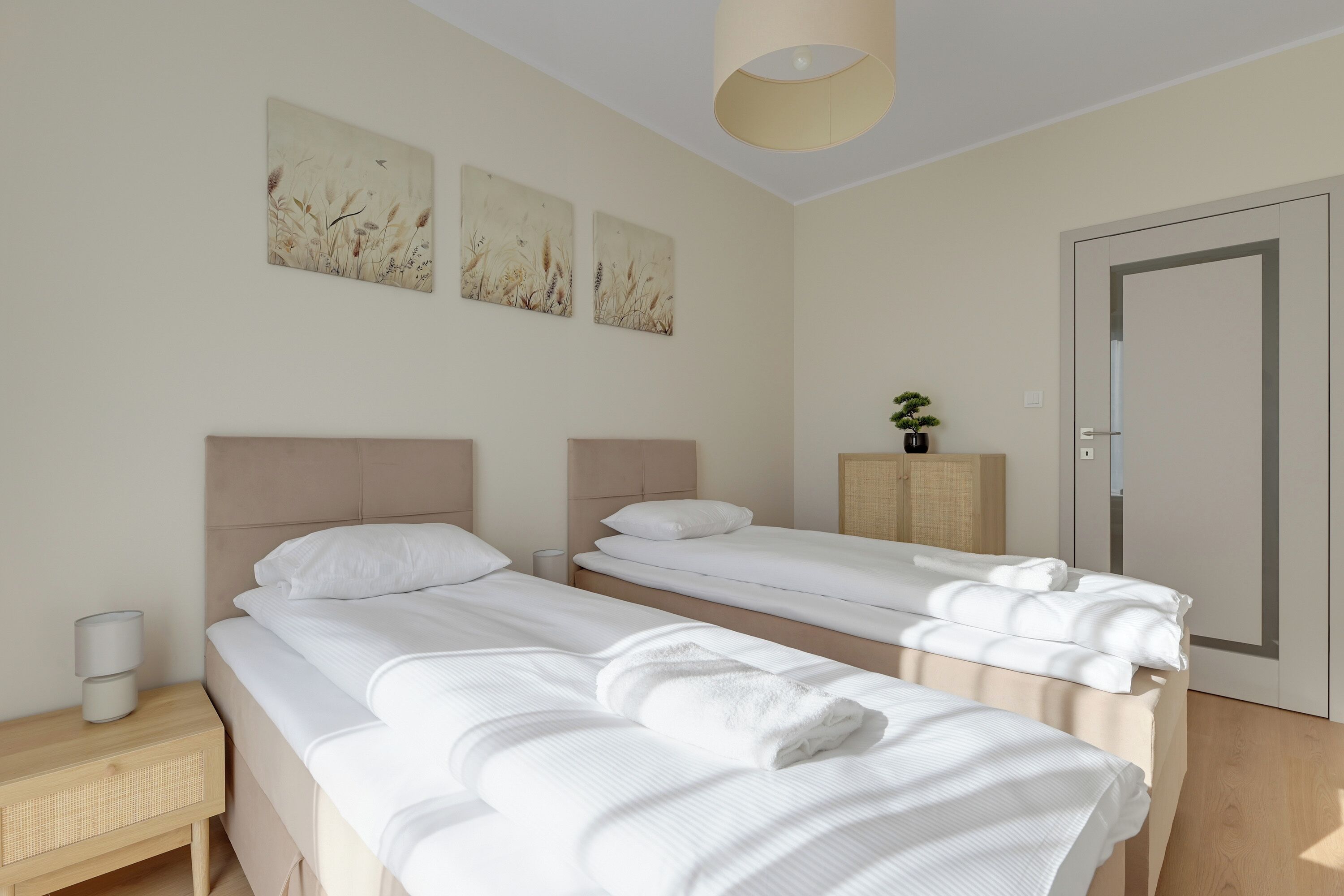 A hotel room with two single beds, a bedside table with a lamp, a small cabinet, and wall art.