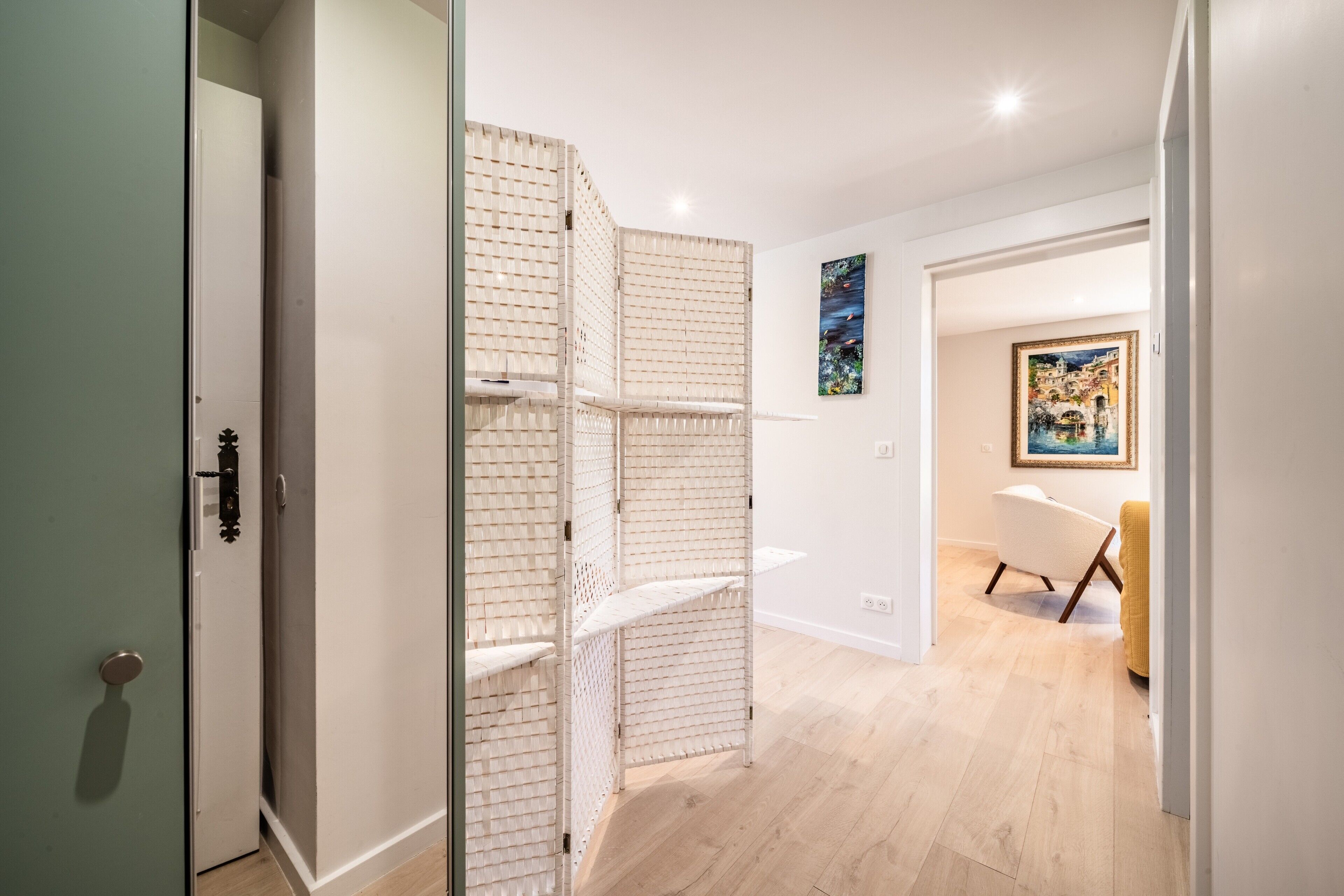 A hallway with a white door, a white partition with shelves, a white chair, and a painting on the wall.