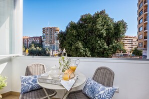 Een balkon met rieten stoelen, een tafel met ontbijtartikelen, en uitzicht op stadsgebouwen en bomen.