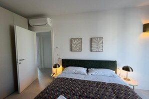 Una camera d'albergo con un letto, due lampade ai comodini, un condizionatore d'aria a parete e due opere d'arte con motivo a foglia.