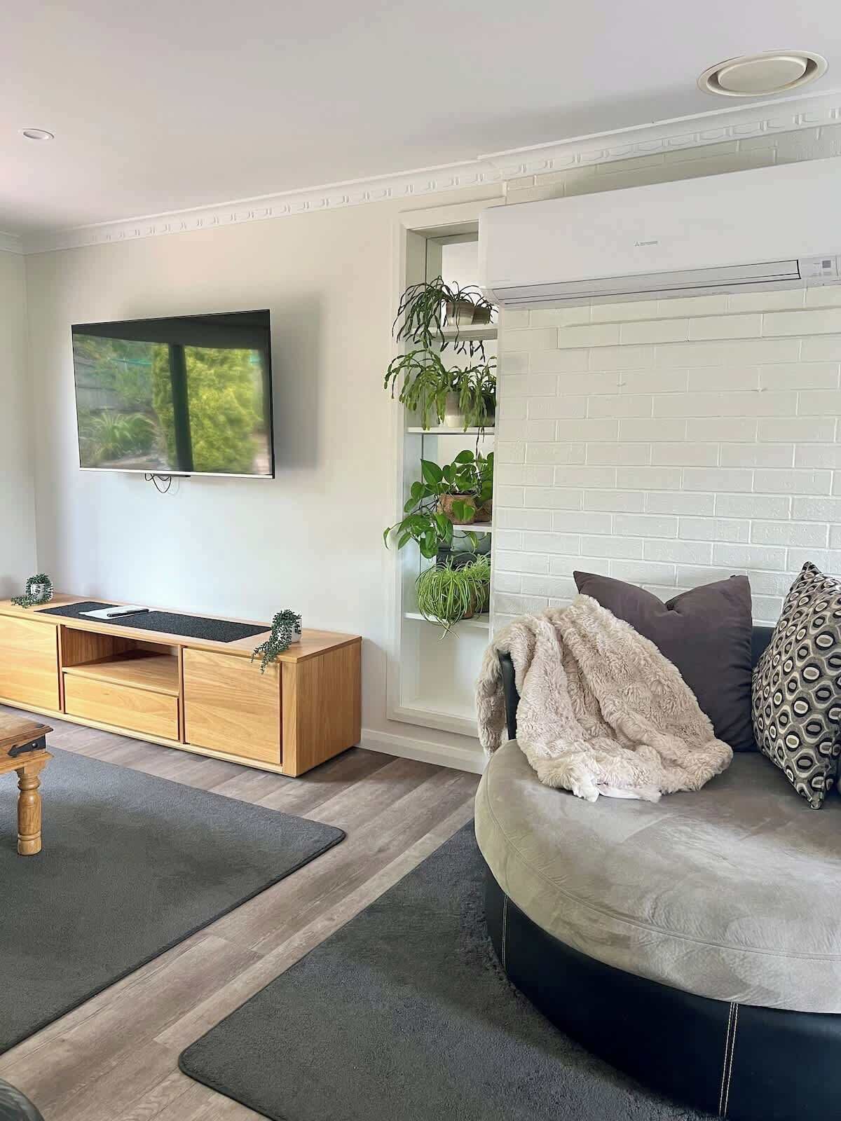 Uma sala de estar moderna com uma televisão de tela plana, um rack de madeira, um sofá redondo e uma estante para plantas.