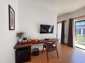 Ein Zimmer mit Schreibtisch aus Holz, Fernseher, Stuhl, kleinem Mülleimer und einem Fenster mit Vorhängen.