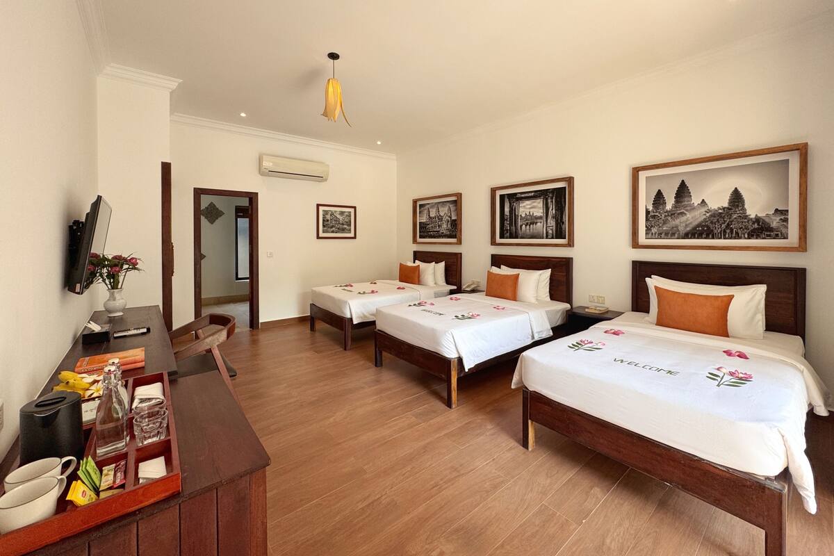 Ein Hotelzimmer mit drei Betten, einem Schreibtisch mit Blumenvase und gerahmten Bildern an den Wänden.