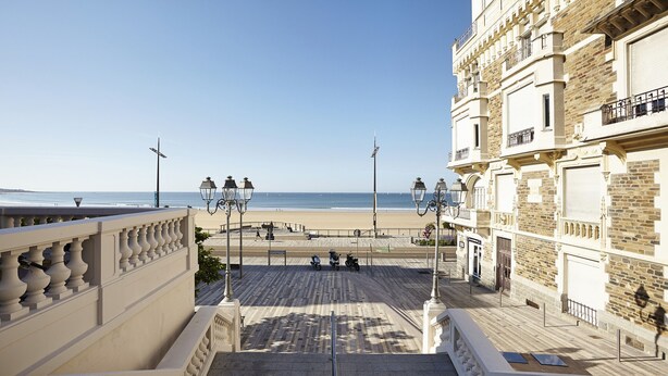 A view from a balcony overlooking a beach, with a building featuring ornate balconies and a paved walkway. - Lovely house with pool for 8 (Les Sables-d´Olonne)