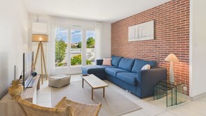 A modern living room with a blue sofa, a glass coffee table, a floor lamp, and a brick wall with a sailboat painting. - Lovely house with pool for 8 (Les Sables-d´Olonne)