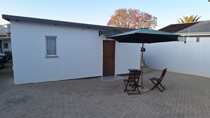 En lille, hvid bygning med en dør og et vindue, en anlagt terrasse med to træstole og et bord, samt en grøn parasol.