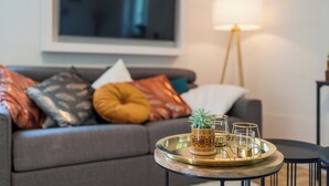 Ein modernes Wohnzimmer mit einem grauen Sofa, das mit bunten Kissen dekoriert ist, einem runden Couchtisch mit einer Schale, in der sich eine Topfpflanze und Gläser befinden, und einer Stehlampe.