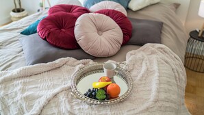 Ein ordentlich bezogenes Bett mit weißer Decke, einem Tablett mit Früchten und einer Tasse sowie bunten Kissen.