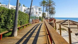 Ein hölzerner Steg führt zu einem Strand mit Palmen und weißen Gebäuden im Hintergrund.