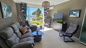 Ein Wohnzimmer mit Sofa, Sessel, Fernseher und Blick ins Freie.