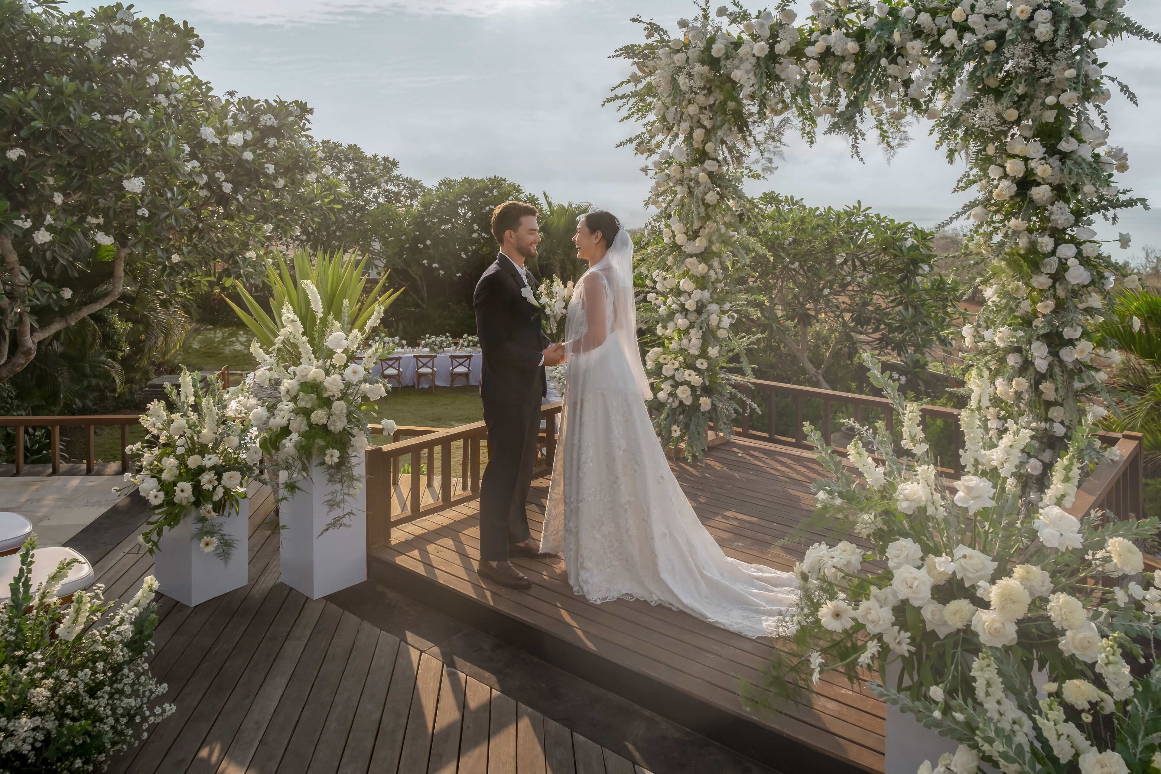 A couple standing under a floral arch on a wooden deck with greenery in the background.