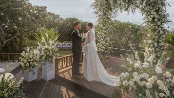 A couple standing under a floral arch on a wooden deck with greenery in the background.