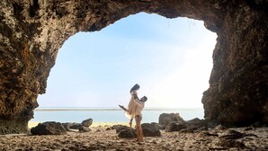A beach scene with a cave entrance, a person in a white shirt, and a clear view of the ocean. - The Shanti Residence By Nakula (Nusa Dua)