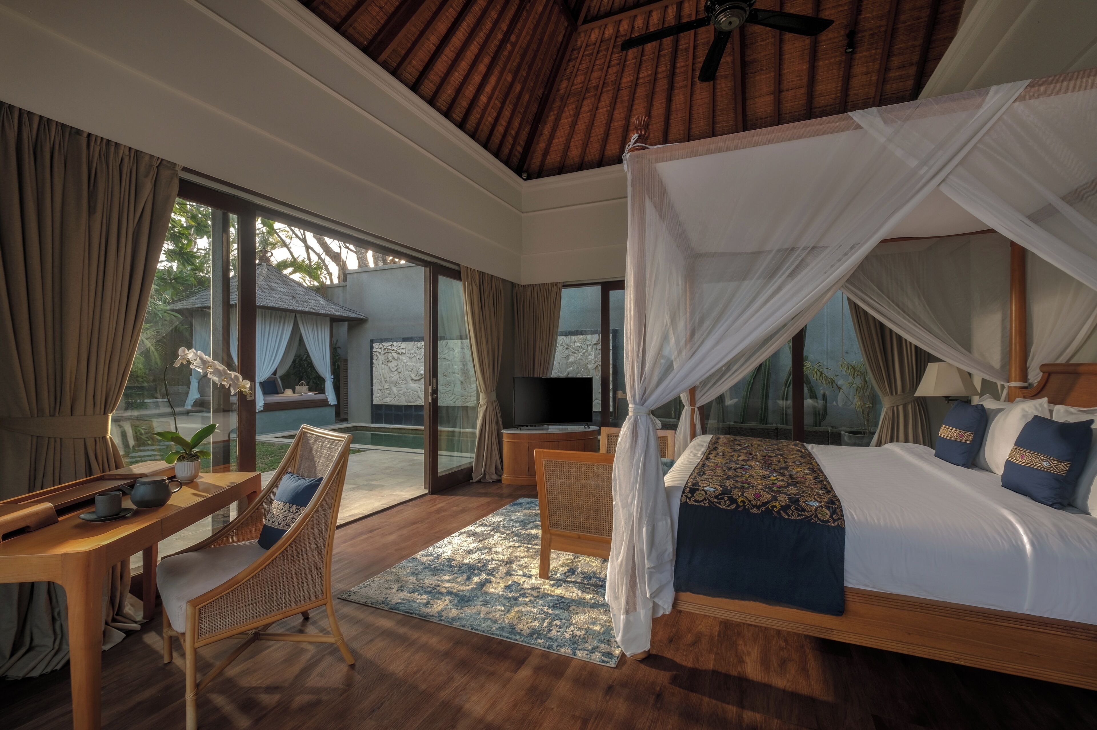 A bedroom with a canopy bed, a television, a desk with a chair, and a view of the outdoors.