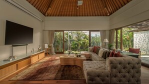 A modern living room with a large flat-screen TV, a wooden ceiling, a large window with a view of greenery, a sofa, and a coffee table. - The Shanti Residence By Nakula (Nusa Dua)