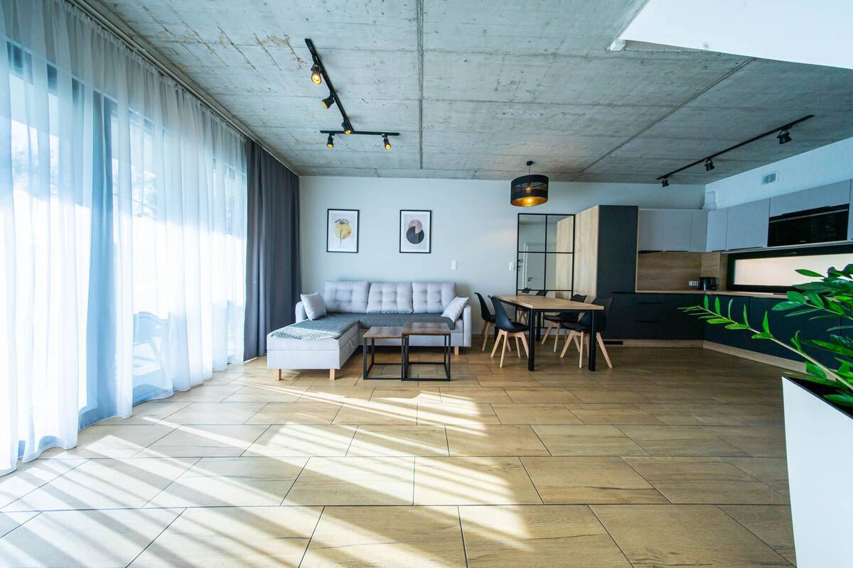 Ein modernes Wohnzimmer mit einer Couch, einem Esstisch, Stühlen, einer Küchenzeile mit Schränken und Geräten sowie großen Fenstern mit transparenten Vorhängen.
