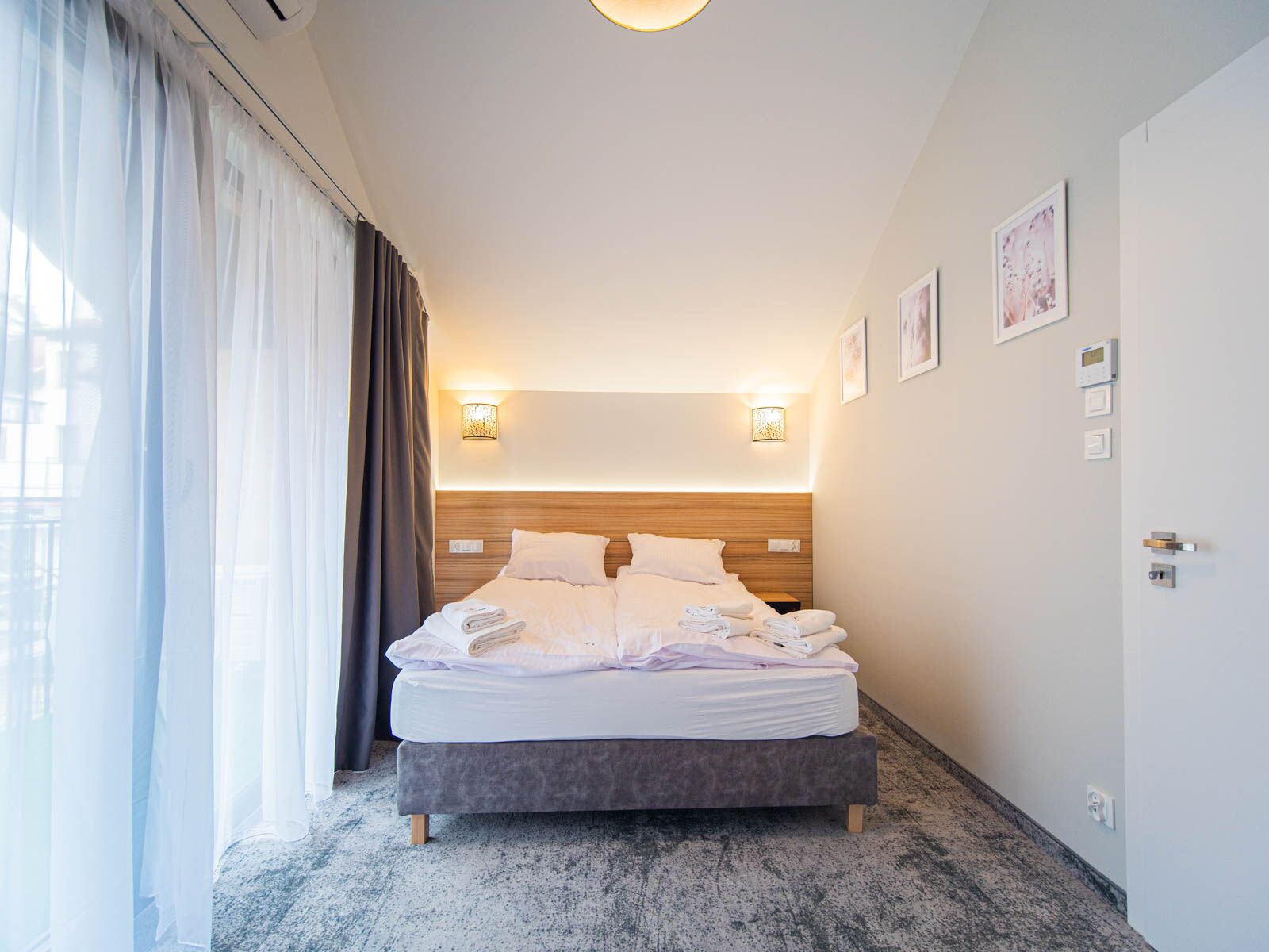 Quarto de hotel com uma cama, duas luzes fixadas na parede, uma porta e uma janela com cortinas.