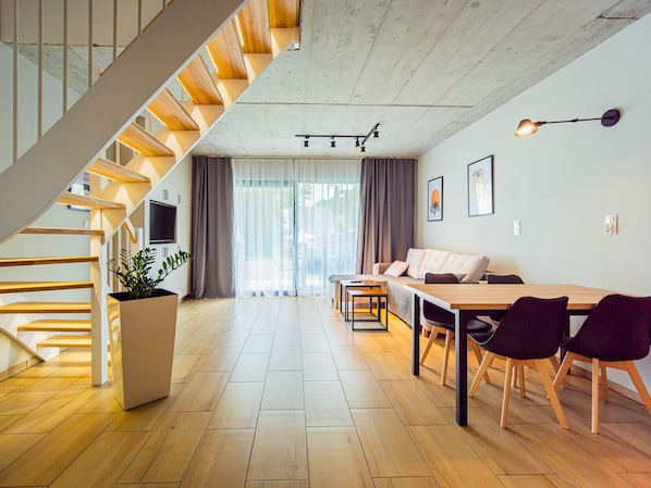 A modern interior with a wooden staircase, a dining area with a table and chairs, a sofa, and a TV mounted on the wall.