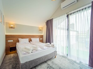 Camera d'albergo con un letto, due luci a parete, un condizionatore d'aria e vista all'esterno attraverso tende leggere.
