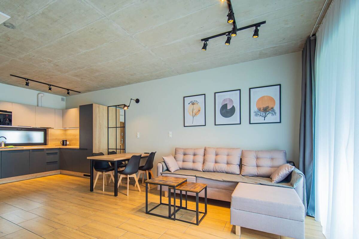 Ein modernes Wohnzimmer mit einer Couch, Hocker und Couchtisch, ein Essbereich mit Stühlen sowie eine Küche mit Arbeitsplatte und Schränken.