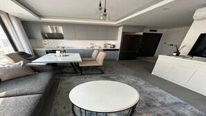 A modern kitchen with grey cabinets, a dining area with a round table, and a sofa. - SOHO (Alanya)