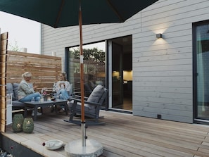 An outdoor seating area with a wooden deck, a covered section, and a modern building with large windows. - 4 Star Holiday Home in Sagad (Sagard)