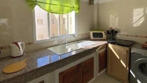 Een keuken met een spoelbak, aanrecht, kastjes, een raam met een groene gordijnroede, een magnetron en een wasmachine.