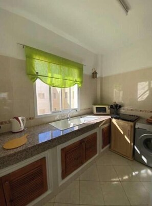 A kitchen with a sink, countertop, cabinets, a window with a green valance, a microwave, and a washing machine. - Charming Le Palmier apartment - Close to the beach, Souk El Had and the town center (Agadir)