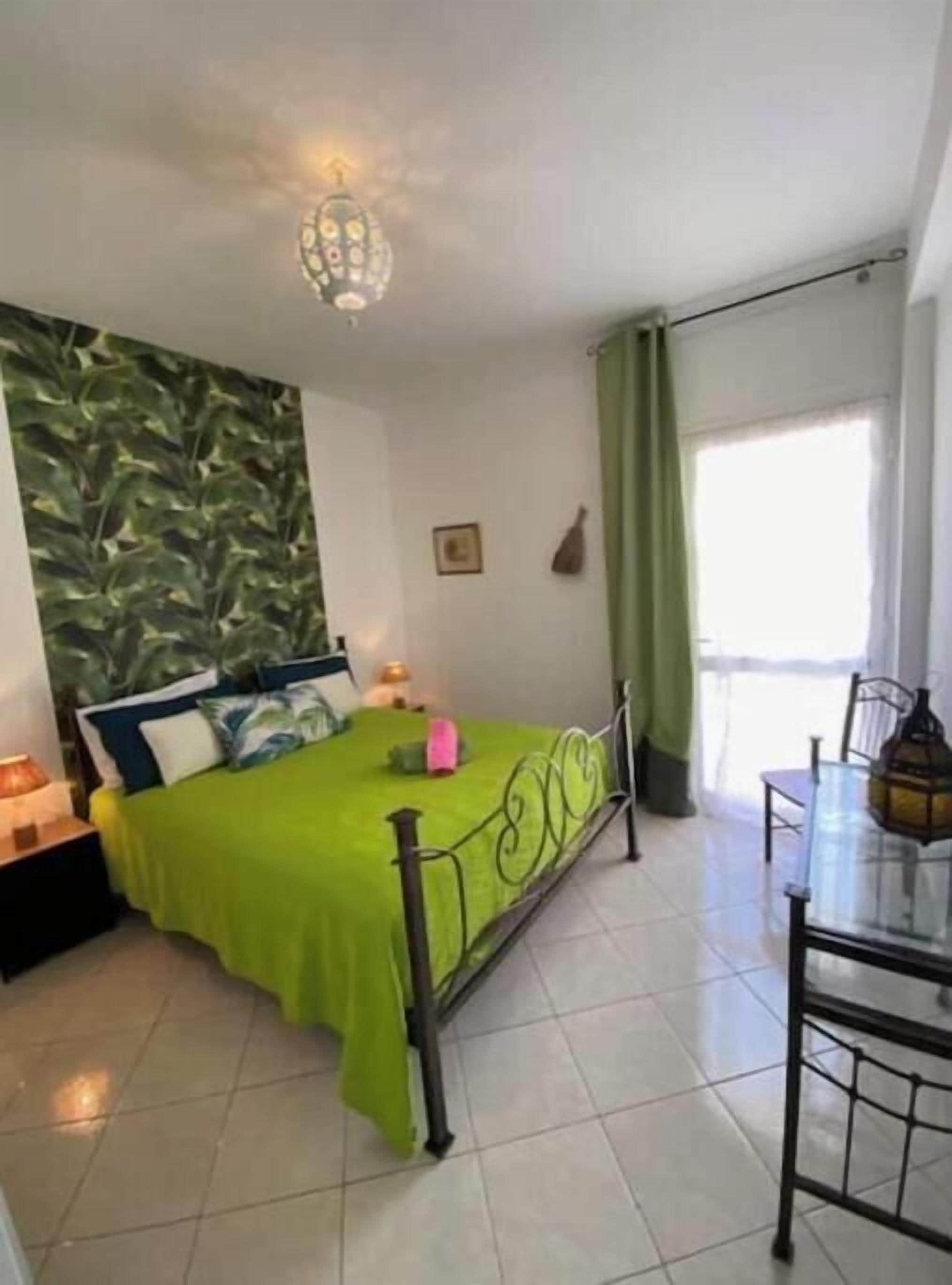 Een kamer met een bed, een tafel, een stoel, een lamp en een raam met gordijnen.