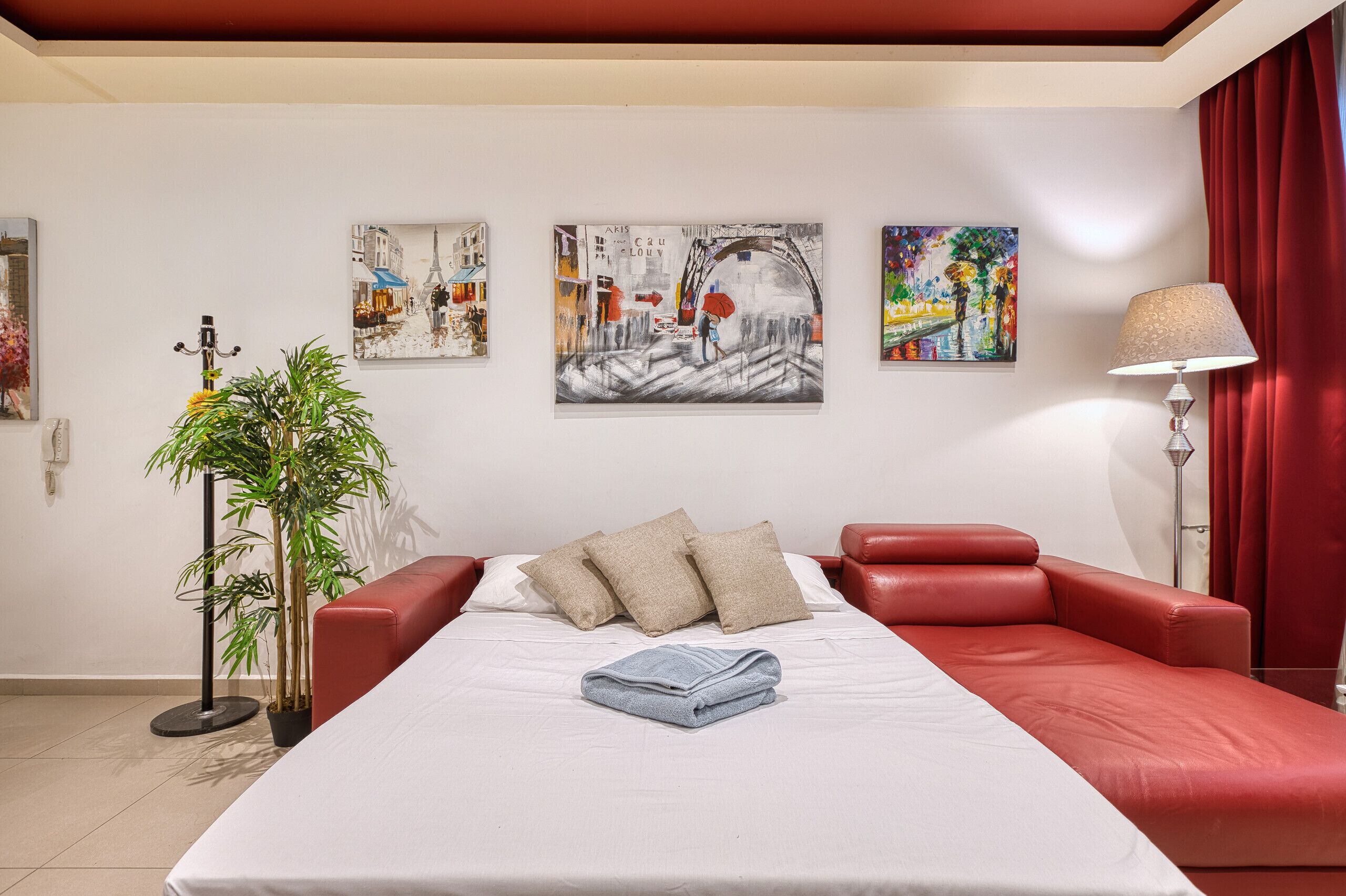 A hotel room with a bed, red leather sofas, a potted plant, and three paintings on the wall.