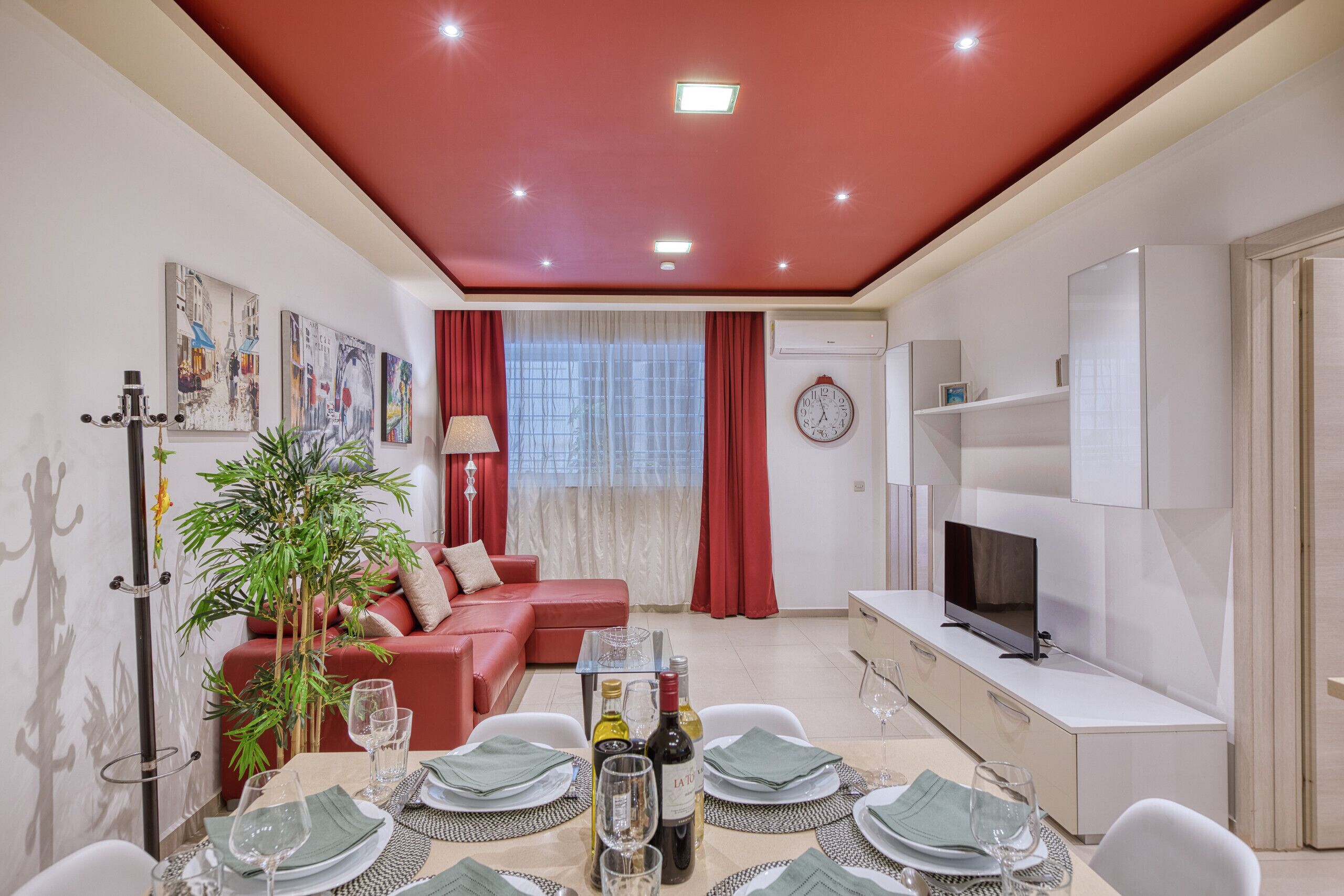 A modern living room with a red ceiling, a sofa, a dining table set for four, and a television on a white cabinet.