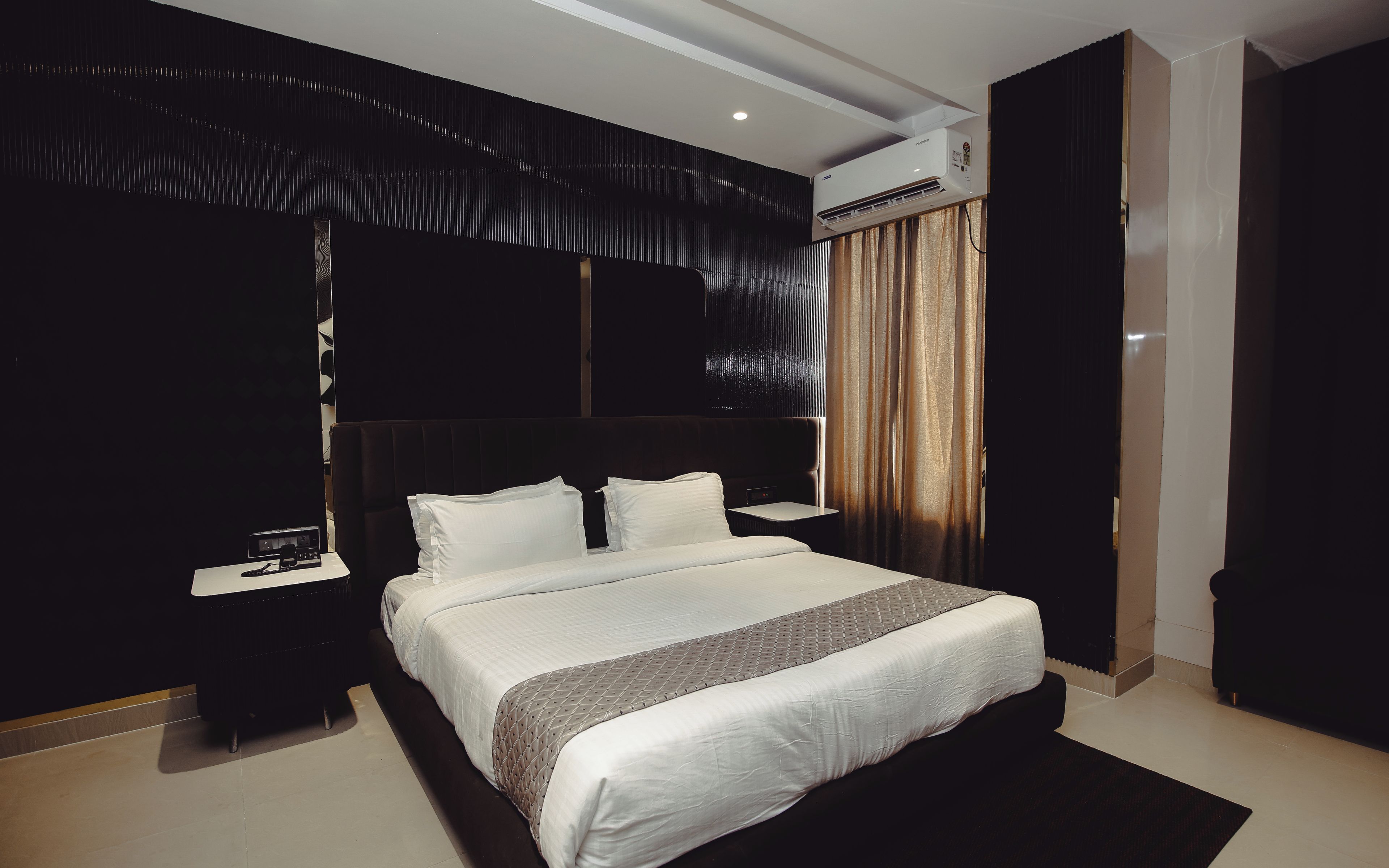 Una habitación de hotel moderna con una cama grande, una mesita de noche, un equipo de aire acondicionado y un cabecero de color oscuro.