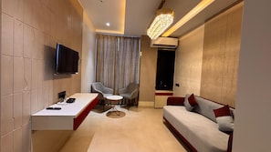 A modern hotel room with a flat-screen TV, a desk with a chair, a sofa, a small round table, and a chandelier. - Dibakar Palace (Tezpur)