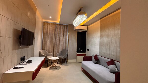 A modern hotel room with a flat-screen TV, a desk with a chair, a sofa, a small round table, and a chandelier. - Dibakar Palace (Tezpur)