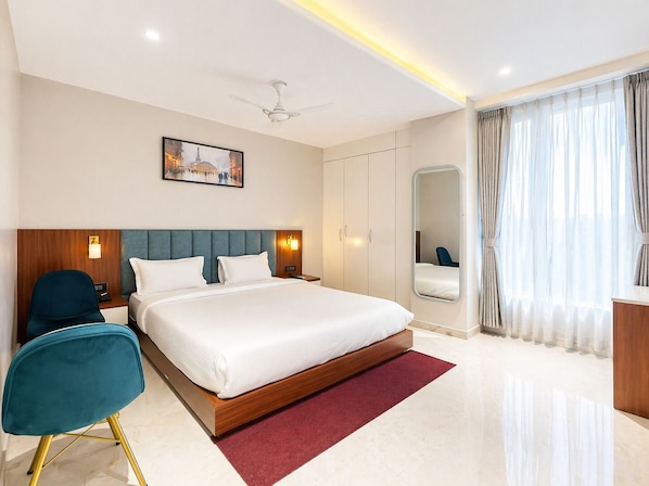 Ein Hotelzimmer mit Bett, Schreibtisch, Stuhl, Deckenventilator, großem Fenster mit Vorhängen, Wandspiegel und einem Gemälde.