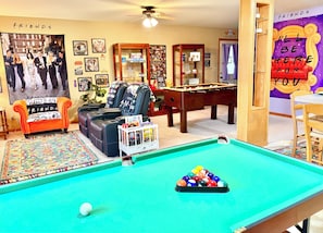 Una mesa de billar con un soporte de bolas colorido, una mesa de fútbol de mesa, un sofá con un cojín con temática de Friends y un cartel de la serie de televisión Friends.