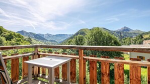 Terraza de madera con mesa metálica, con vista a un paisaje montañoso de abundante vegetación y cumbres lejanas.