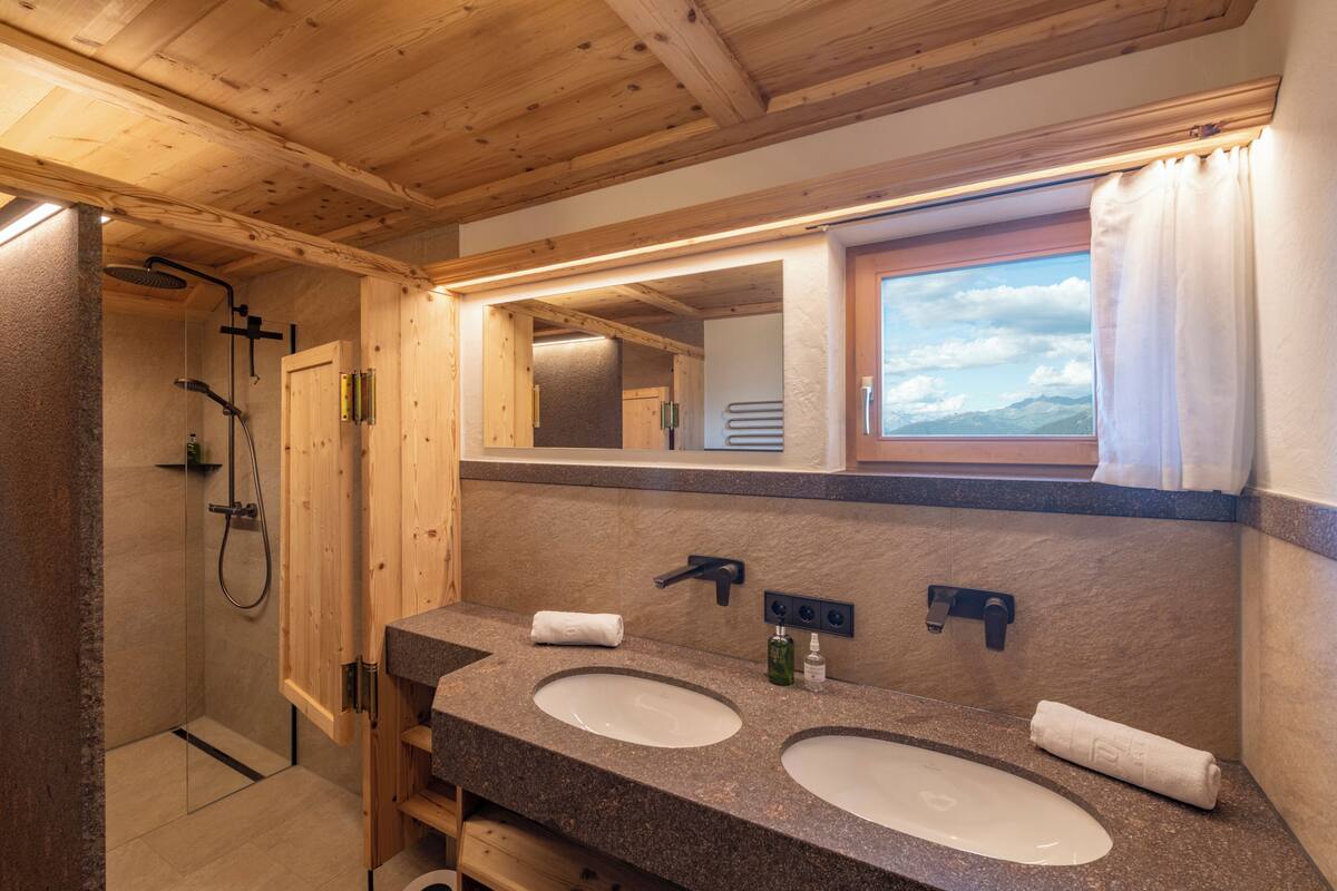 Une salle de bain avec des poutres en bois, deux lavabos, une douche et une fenêtre donnant sur un beau paysage.