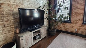 Ein Zimmer mit Backsteinwand, einer Holz-Unterhaltungsecke mit Flachbildfernseher, einer Topfpflanze und einem Fenster mit Jalousien.