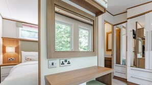 A modern hotel room with a bed, bedside table, lamp, mirror, and built-in wardrobe. - Fern View - Kelling Heath Holiday Park (Holt)